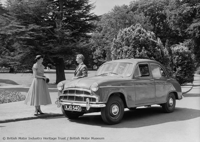 A black and white photograph of a Morris Isis Saloon parked at the side of the road near a park. A man wearing a suit rests his arm on the car's bonnet as he faces a smiling woman wearing a dress and a hat. 