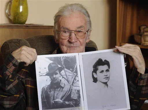An older man holding two photographs, one of a young individual wearing a hat (himself) and the other of a woman with styled hair.

