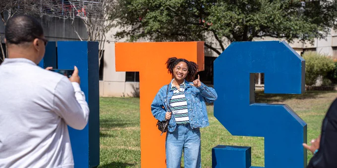 A person posing with a peace sign in front of large orange and blue letter sculptures displaying "UTSA," while another person takes their photo. They are outdoors on a sunny day with trees and a building in the background.