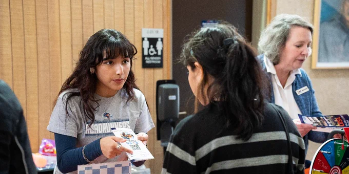 Person handing another person a brochure at a community event, with a third person standing nearby, all in a room marked with a restroom sign indicating accessibility.