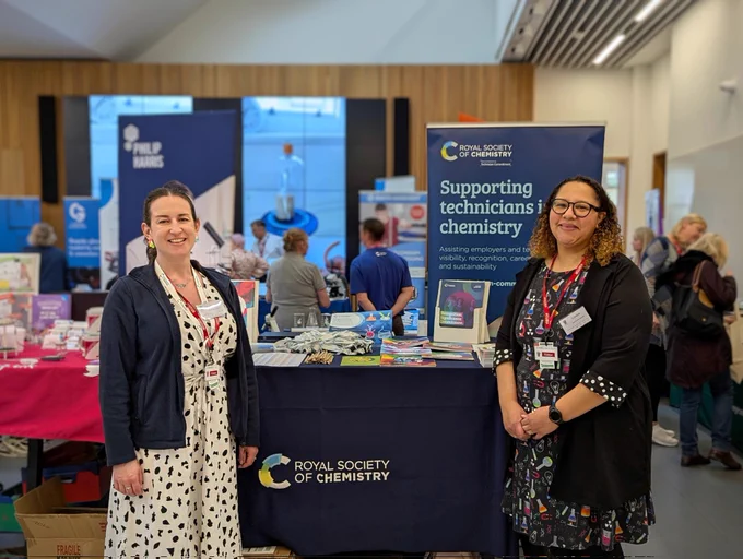 Two women standing in front of a Royal Society of Chemistry exhibition stand, with a banner behind with the text "supporting technicians in chemistry". People are milling about around the room.