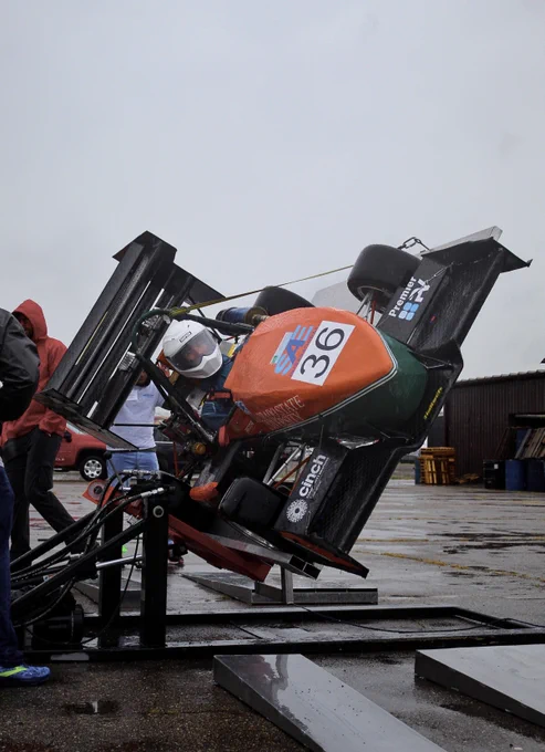 A Formula SAE car lifted into the air and tilted to the side.  A driver sits in the car.