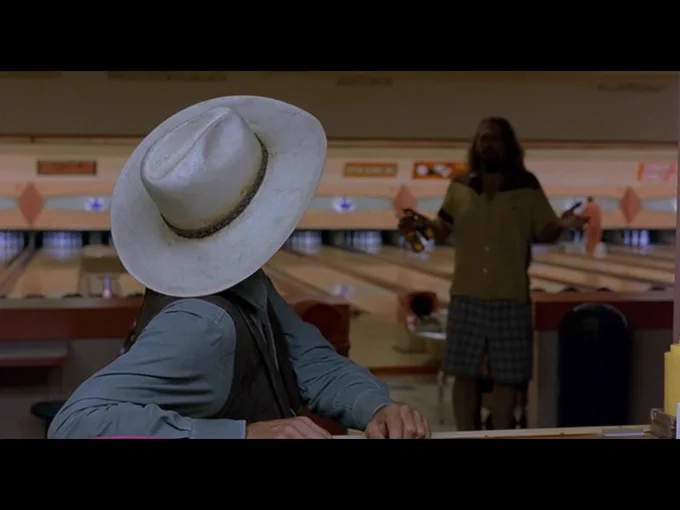 In the background, the Dude turns to face the Stranger at the bar one last time.