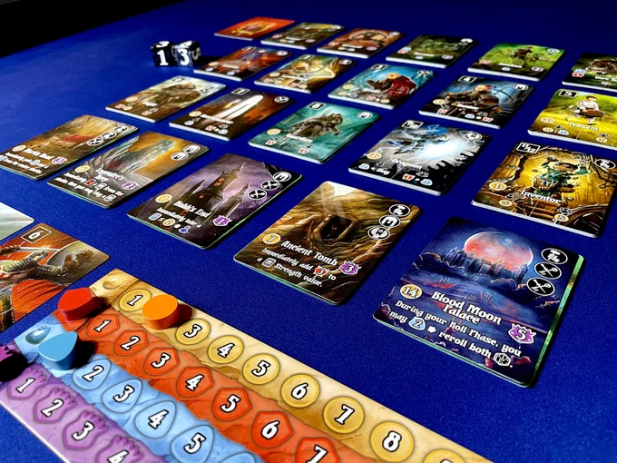 The game Valeria Card Kingdoms set up on a table with cards from the Shadowvale expansion. There is a player board beneath a grid of card stacks and dice in the background.