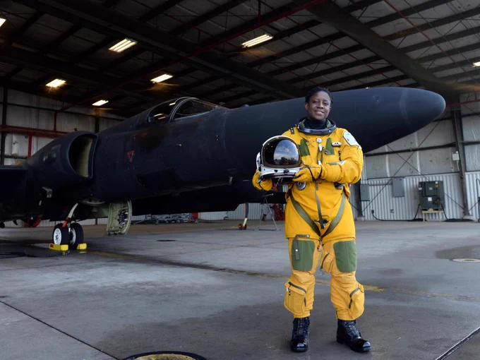 A woman in a yellow flight suit stands in front of a black U-2 aircraft inside a hangar. The pilot is holding a helmet and smiling at the camera.