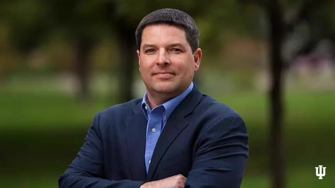 Person in a blue blazer standing outdoors with blurred greenery in the background and the Indiana University logo visible in the corner.