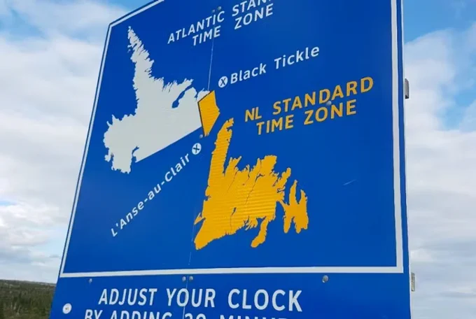 The photo shows a blue highway sign indicating a time zone change between the Atlantic Time Zone and the Newfoundland and Labrador (NL) Standard Time Zone. The sign features a map highlighting Newfoundland in yellow and Labrador in white, with locations like Black Tickle and L’Anse-au-Clair marked. Text on the sign reads "ADJUST YOUR CLOCK BY ADDING 30 MIN." The background shows a cloudy sky.