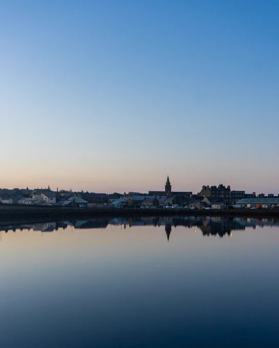 View of Kirkwall silhouetted against a blue and pink sunrise, with the calm waters of Kirkwall bay in the foreground. 