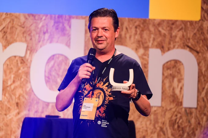 Man on stage holding a microphone and a 'U' shaped award, wearing a black t-shirt with a graphic design, against a backdrop with rustic wood panels.