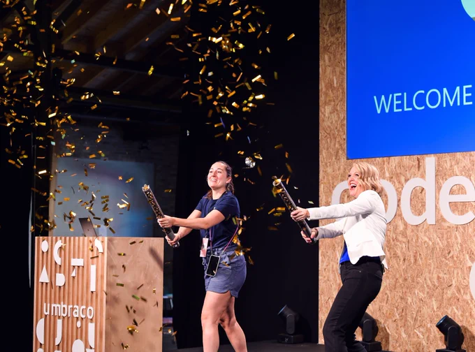 Two women on stage, one in a blue t-shirt and shorts, the other in a white blazer and black pants, joyfully shooting confetti cannons, with golden confetti in the air and a 'Welcome' sign on a screen.