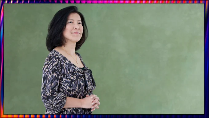 A portrait of Yoko Shimomura . She wears a patterned blue and white dress, and thoughtfully holds her hands in front of her as she looks out to the distance. She stands in front of a textured green backdrop.