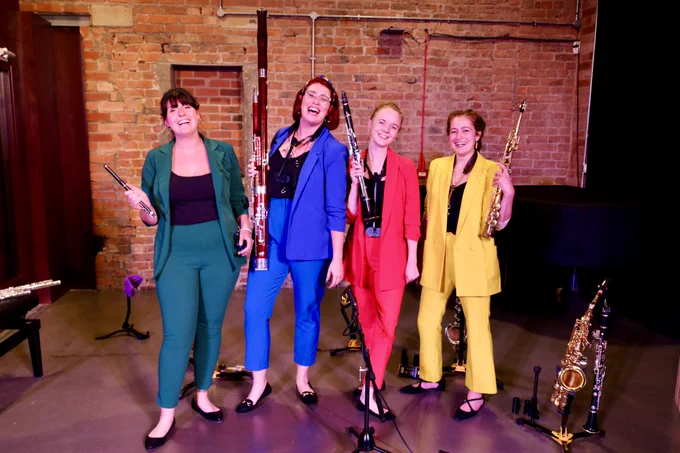 RPS Henderson Chamber Ensemble award winners Chameleon Quartet against a red brick background, each wearing a different coloured suit (green, blue, red and yellow), and holding their instruments (piccolo, bassoon, clarinet, and soprano saxophone) with more instruments on stands in the background  (saxophones, clarinet, flute).