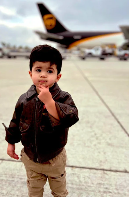 Child in a brown leather jacket on an airport tarmac with a UPS plane in the background.