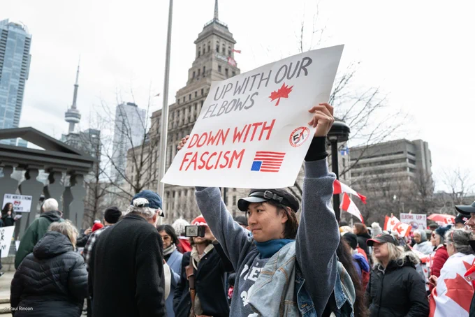 Person holding up a sign at a demonstration "UP WITH OUR ELBOWS (RED MAPLE LEAF) DOWN WITH FASCISM (Upside down US flag - no 51).