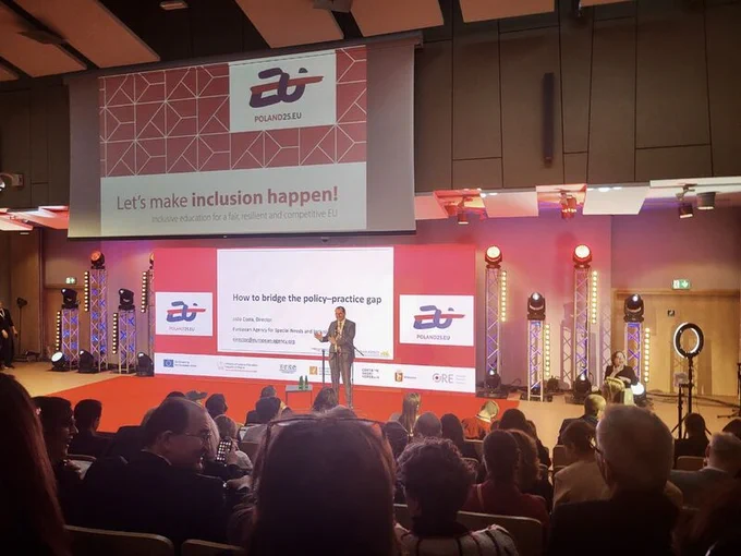 picture of the plenary of the conference - A man speaks into a microphone at a conference, addressing an audience seated in front of him. behind him the first slide of a powerpoint presentation, stating "Let's make inclusion happen!"