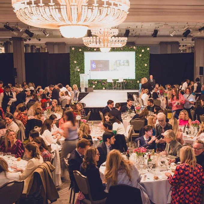 Guests seated at round tables during a banquet event in a large hall with a central chandelier and a stage displaying a presentation.
