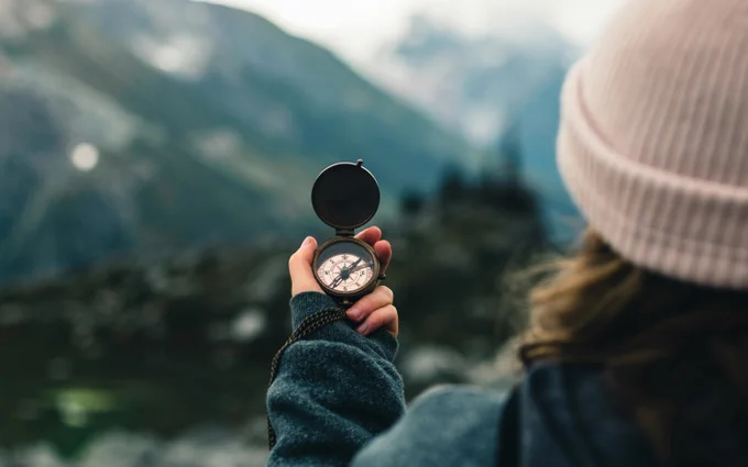 A view from the rear of woman with a white knit knit cap holding a compass with mountains before her. (Photo Credit: Ali Kazal on Unsplash)