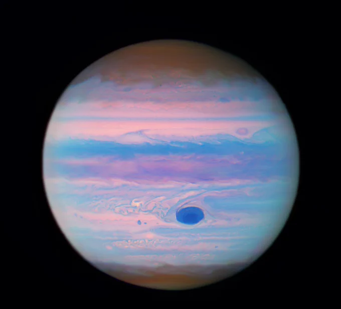 The planet Jupiter with its cloud bands seen in shades of pink, rusty red, blue, and purple. The Great Red Spot is a deep navy blue, surrounded by bands and swirls of pink, and light blue.