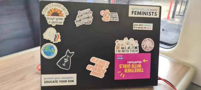 Black Laptop on a table, with a series of stickers on it such as a Plan International UK logo, a sunflower with the slogan today is a food day to disrupt systems of oppression, a small ghost with an lgbt rainbow flag, a planet earth with the slogan destroy the patriarchy not the planet, a black dress with the slogan it's a dress not a yes, text of protect your daughter crossed out with educate your son in bolder text underneath, a sticker of the slogan old ways won't open new doors, text saying fueled by caffeine and Feminist rage surrounded by flowers, a cat with a stop sign saying negative self talk forbidden in this area, text saying boys will be boys with the second boys crossed out to read boys will be feminists, cats paws and hearts saying I'm with them, and cats against the patriarchy, and text sating you are human its OK to feel. 