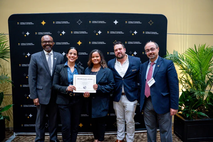 Dean Grant Hayes and Associate Dean Naim Kapucu standing and smiling in front of a UCF College of Community Innovation and Education backdrop with the UCF students who placed second in the Research Impact Showcase.