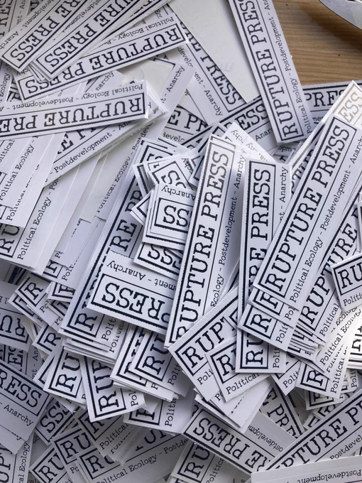 smaller rectangular stickers with the text RUPTURE PRESS, and the words political ecology- postdevelopment- anarchy underneath. The stickers are stacked on top of each other on a wooden table