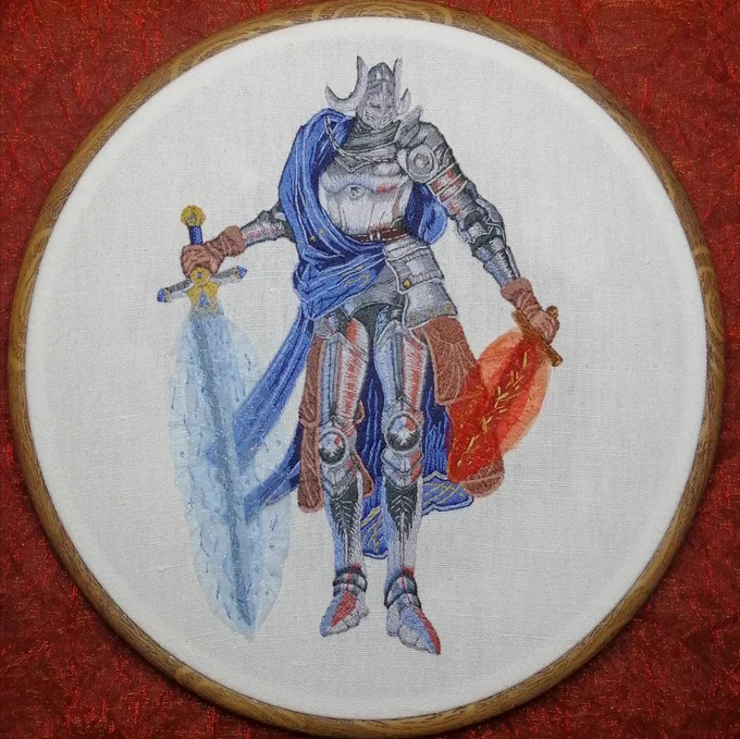 Wooden embroidery hoop on a red organza background: the subject is Rellana, a knight in full armor with two moons on the sides of her helmet, two swords and a cape draped over her right shoulder. The canvas is white. The helmet, torso, arms and legs are embroidered with several shades of grey, light blue highlights on the left and red highlights on the right. The belt and the gloves are brown and the cape is embroidered in shades of blue with golden details. The sword on the left is shrouded in blue organza, the one on the right in red. 