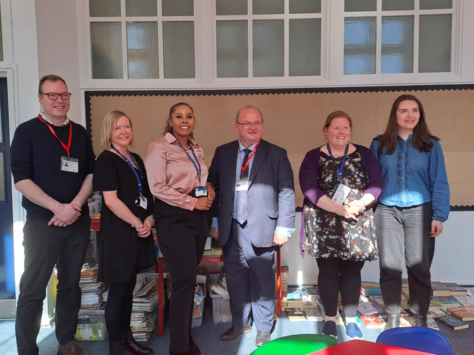 Six people standing in front of a room with books scattered on the floor, awaiting its transformation into a library.