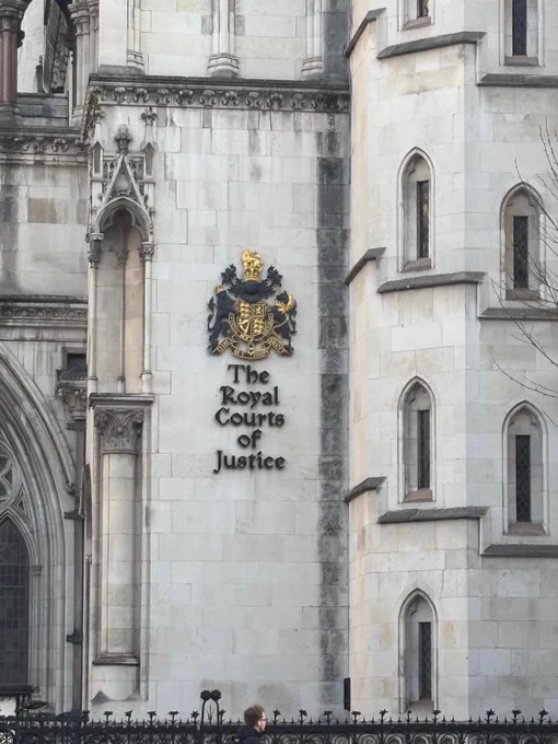 The exterior of the Royal Courts of Justice in London. The stone facade has the UK royal coat of arms above the black text "The Royal Courts of Justice." The architecture features pointed arch windows, decorative carvings, and stone detailing. A person is walking by outside the wrought iron fence at the bottom of the image.