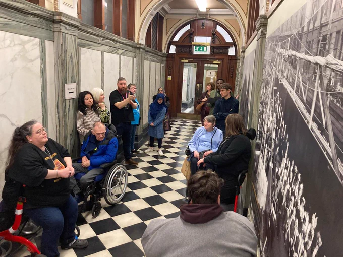 A group of GDA members in a hallway with chequered floors. They are listening to a Mitchell Library tour guide.
