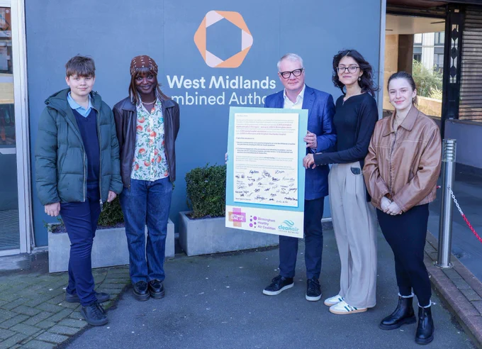 Five people standing outside of a building, with one person holding a giant signed letter.