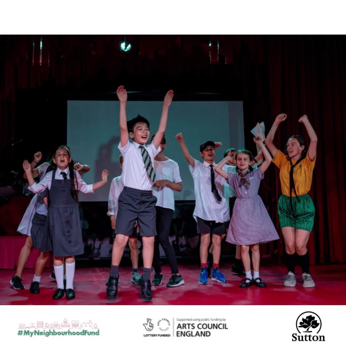 A group of children on stage, mid-performance, with joyful and energetic expressions. They are dressed in various costumes, including school uniforms and colorful outfits. One boy in the center is jumping with his arms raised high, while others around him cheer and pose with their arms in the air. The stage has a red floor, and a large screen in the background adds to the theatrical atmosphere. Curtains frame the sides of the stage, enhancing the performance setting.