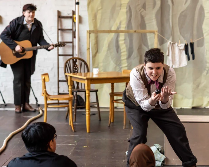 Two children look up at an actor, who crouches to show them a large marble. In the background, another actor plays a guitar.