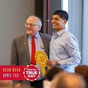 Two individuals standing together at an event, one wearing a suit and the other in a dress shirt, smiling with a "Forever True" ribbon visible. The setting appears to be a formal gathering or ceremony.