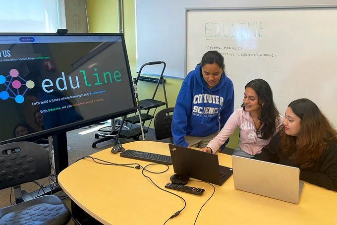 Three students look at a laptop screen together. At left is a large screen that says eduline, the name of the generative AI project they developed  that will help preserve languages in the future. 