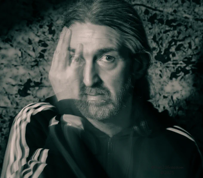 This self-portrait captures a person wearing a dark jacket with white stripes. Through the use of slow exposure, the image features a subtle motion blur from the subject's hand, while one eye, partially obscured by a finger, still remains in focus, creating an intriguing, somewhat surreal effect. The composition is set against a textured, abstract monochrome backdrop, enhanced by dramatic lighting that highlights the contrast between clarity and movement within the frame.