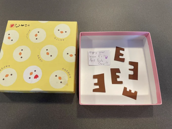 Open gift box with a handwritten note that says "Enjoy your brown E’s! April Fools Day!!" accompanied by several cut-out letter "E"s made of brown paper. The box exterior features a cute yellow pattern with chick faces and Japanese text. This is a humorous April Fools’ prank playing on the expectation of receiving brownies.