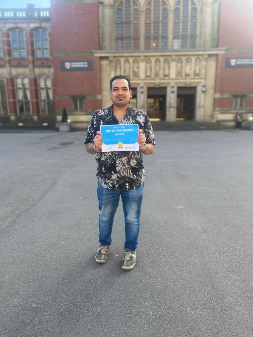 Image of a student in front of Aston Webb smiling at the camera and holding a certificate for Rep of the Month.