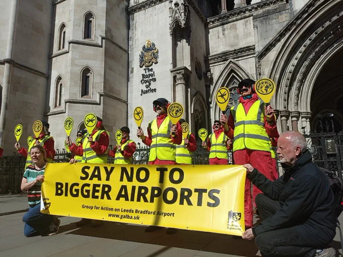 A group of people in high vis holding airplane landing signals with a no planes icon and a world in flames icon, standing in front of the Royal Courts of Justice in London. Two people are holding a yellow banner in front which says 'say no to bigger airports'.