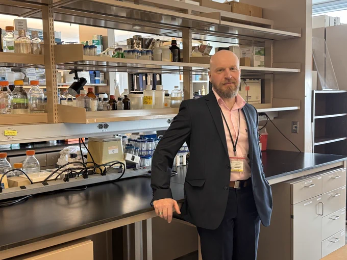 Photo of a man in collard shirt and jacket, standing in a research lab. 