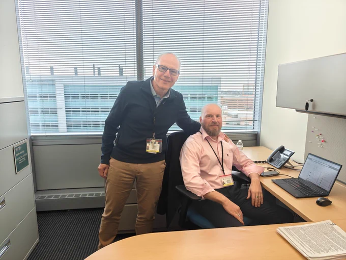 Photo of two men in collard shirts. One standing one sitting in an office. 