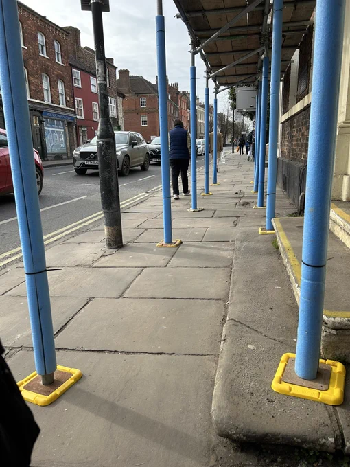 Blue scaffolding with stone step jutting out into the clear width with a lamp post on the outside of it that makes the pavement completely impassable to many disabled people.