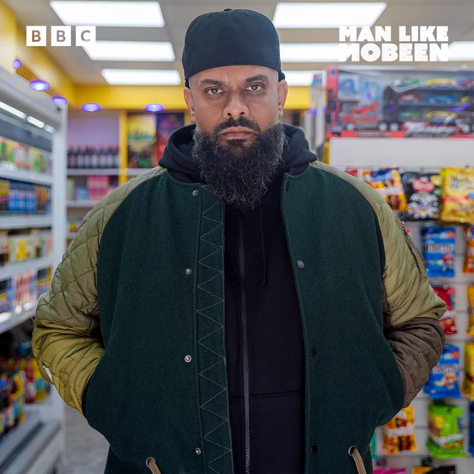 Mobeen in a dark hoodie and green bomber-style jacket stands in the aisle of a convenience store. He has a serious expression.
