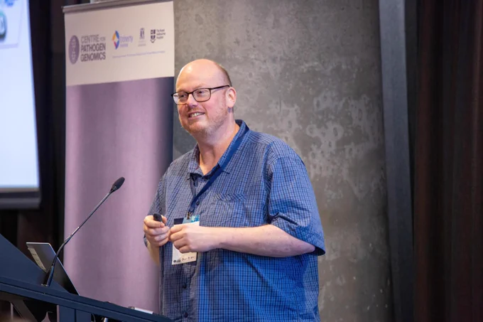 This image shows a middle-aged man with glasses and a beard standing at a podium, delivering a presentation. He is wearing a blue checkered short-sleeve shirt and a lanyard with a name tag. He holds a presentation remote in his hands and is smiling while speaking.

Behind him, a banner displays the logo and name of the Centre for Pathogen Genomics, along with additional institutional logos, suggesting a scientific or academic conference setting. The background consists of a textured gray wall, and a microphone is positioned near the speaker, indicating a professional speaking environment. The image captures a moment of engagement, where the speaker appears to be explaining or presenting research findings to an audience.