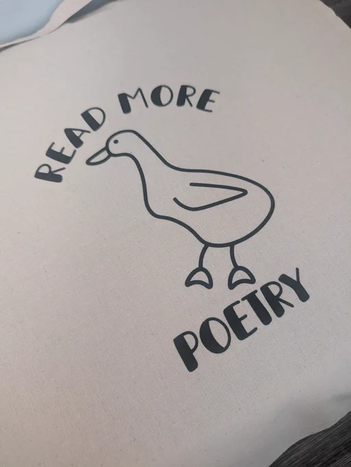 A close-up photo of a tote bag. Printed in black on the bag is a simple line drawing of a duck, with the words "READ MORE" above it and "POETRY" below it, arranged in a playful curved style.