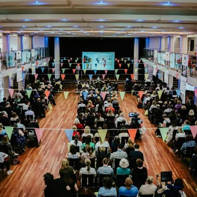 An audience seen from above, from a mezzanine floor. All facing towards a large screen. Colourful bunting is hung above their heads.
