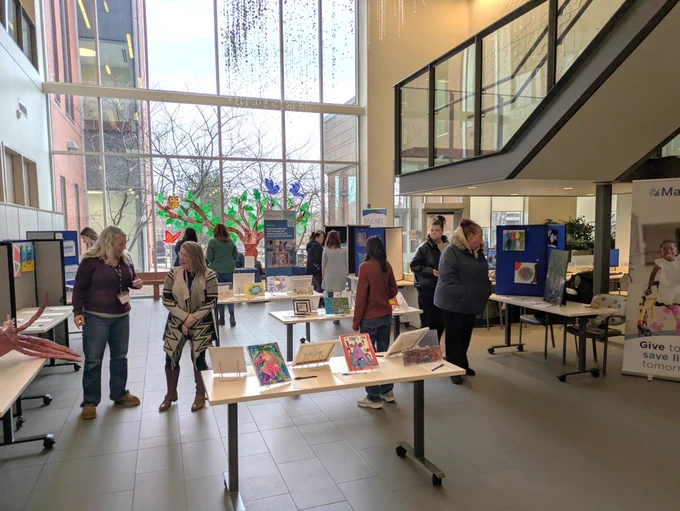 A bright auditorium set up with tables featuring artwork created by Autistic children and youth. Viewers are walking around enjoying the art and conversing.