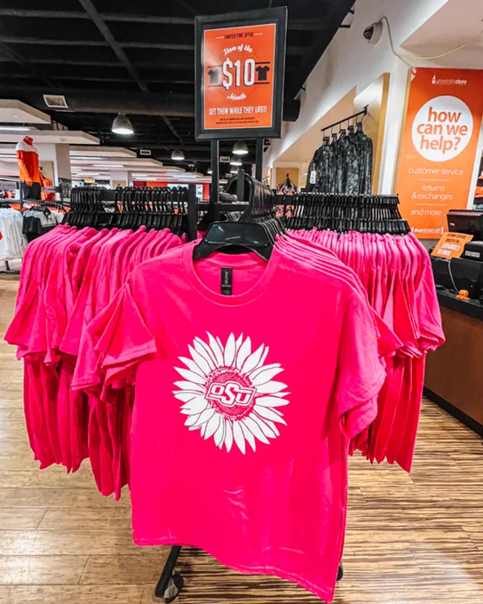 Rack of bright pink t-shirts with the Oklahoma State University logo displayed in white at the center of a sunflower. 