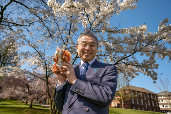 A man in a suit smiles, holding a squirrel teddy, in front of a cherry blossom tree with a blue sky in the background.