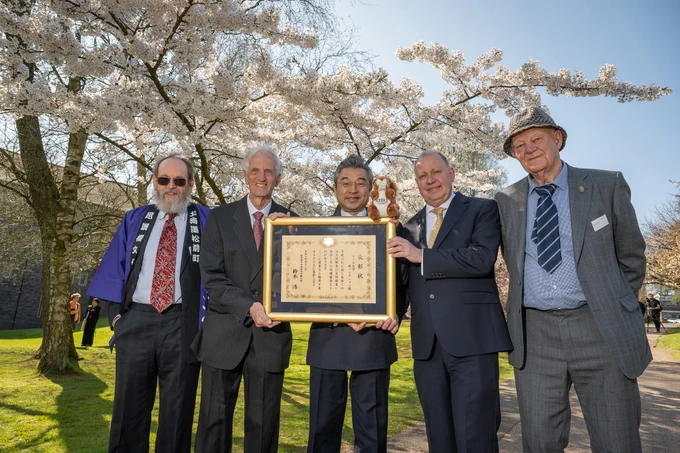 A group of people stand in front of cherry blossom trees with white flowers, holding a certificate in a frame and a squirrel teddy.