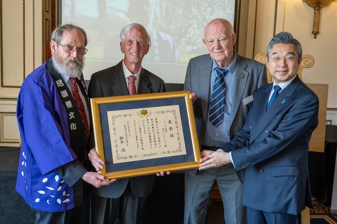 Four people smile holding a certificate in a wooden frame, in front of a projector screen.